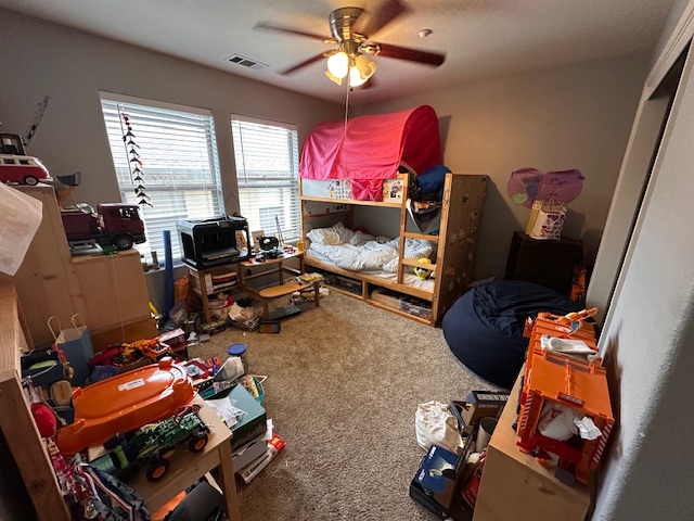 Spacious kid’s room with bunk bed, carpeted floor, large windows, ceiling fan, and shelves filled with toys and books.