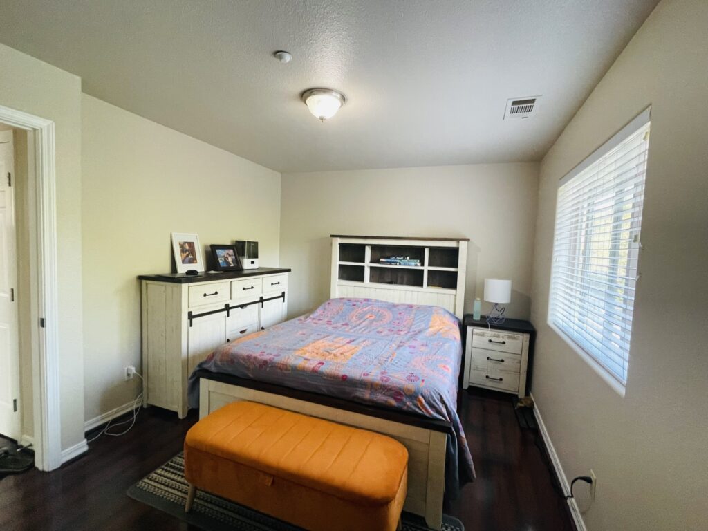 Alternate angle of primary bedroom showing dresser, nightstand, and window with blinds.